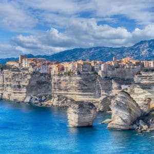 Una città costiera con edifici tradizionali si erge su alte scogliere bianche, affacciata su un mare azzurro, con montagne sullo sfondo.