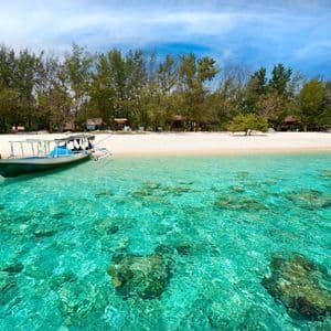 Ein Boot schwimmt in flachem, türkisblauem Wasser, das den felsigen Meeresboden durchscheinen lässt, nahe einem bewaldeten Sandstrand.