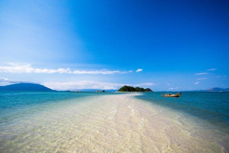 Un banco de arena blanca se curva a través de aguas claras y poco profundas hacia una isla verde distante, con un pequeño bote flotando cerca.