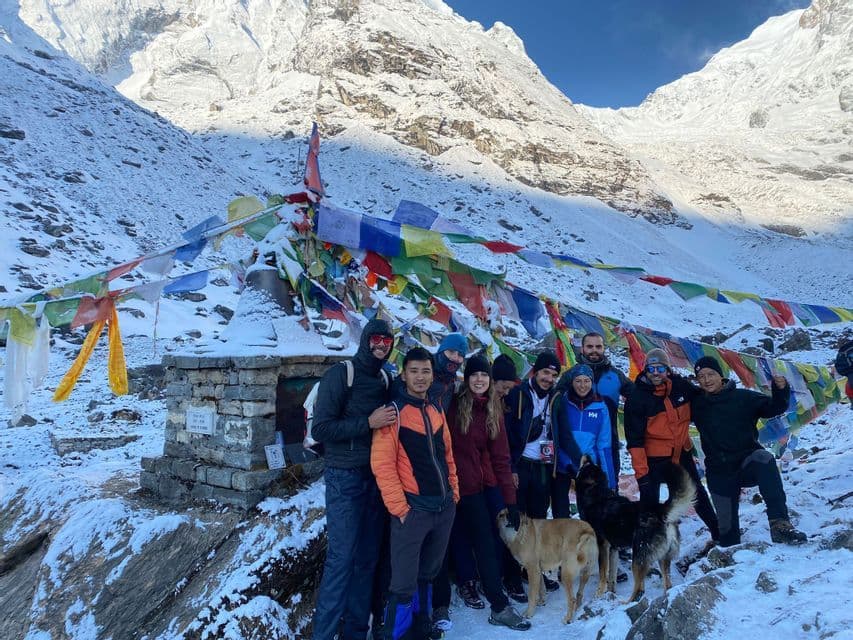 Un voyage de groupe WeRoad avec deux chiens posant pour une photo devant un sanctuaire avec des drapeaux de prière colorés dans une chaîne de montagnes enneigée.
