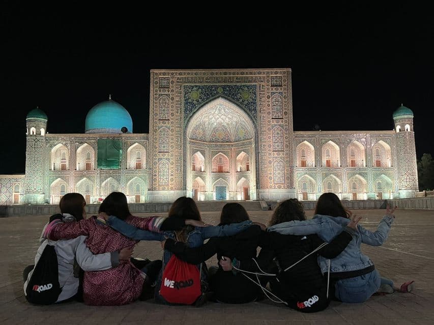 Seis mujeres de un viaje en grupo de WeRoad están sentadas en el suelo con los brazos entrelazados, frente a un edificio ornamentado e iluminado de noche.
