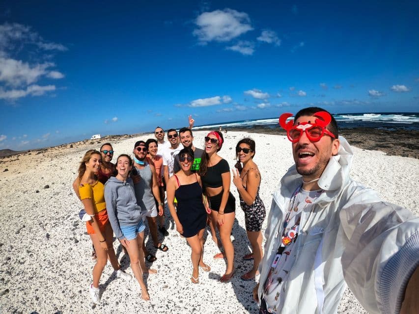 Un homme avec des lunettes rigolotes en forme de crabe prend un selfie grand-angle de son voyage de groupe WeRoad sur une plage de galets blancs.