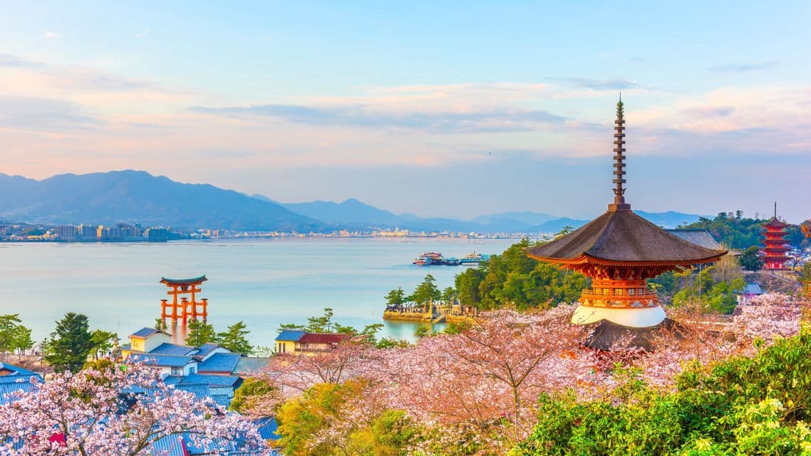 Ein malerischer Blick auf eine japanische Pagode und ein Torii-Tor am Wasser, umrahmt von Kirschblüten, mit Bergen im Hintergrund.