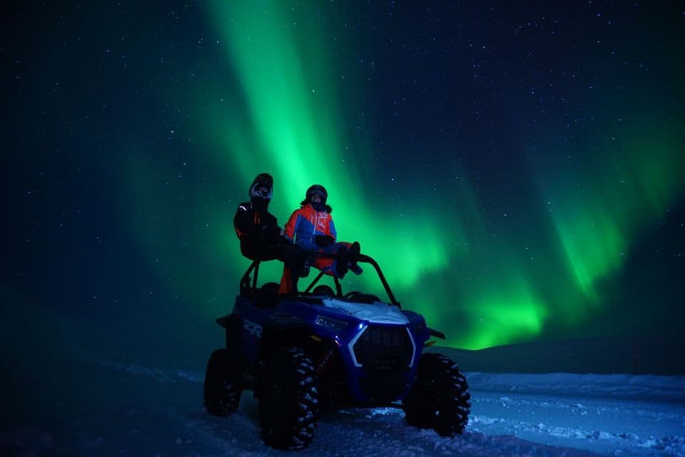 Zwei Personen mit Helmen sitzen auf einem Geländefahrzeug im Schnee und beobachten die Nordlichter, die den sternenklaren Nachthimmel erhellen.