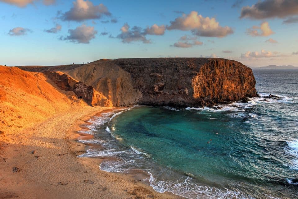 Una spiaggia sabbiosa si estende in una baia con acque turchesi, fiancheggiata da scogliere rocciose illuminate da una luce dorata sotto un cielo parzialmente nuvoloso.