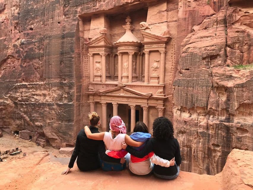 Un grupo de viaje de WeRoad de cuatro mujeres sentadas abrazadas, admirando un antiguo templo tallado en un acantilado de roca.