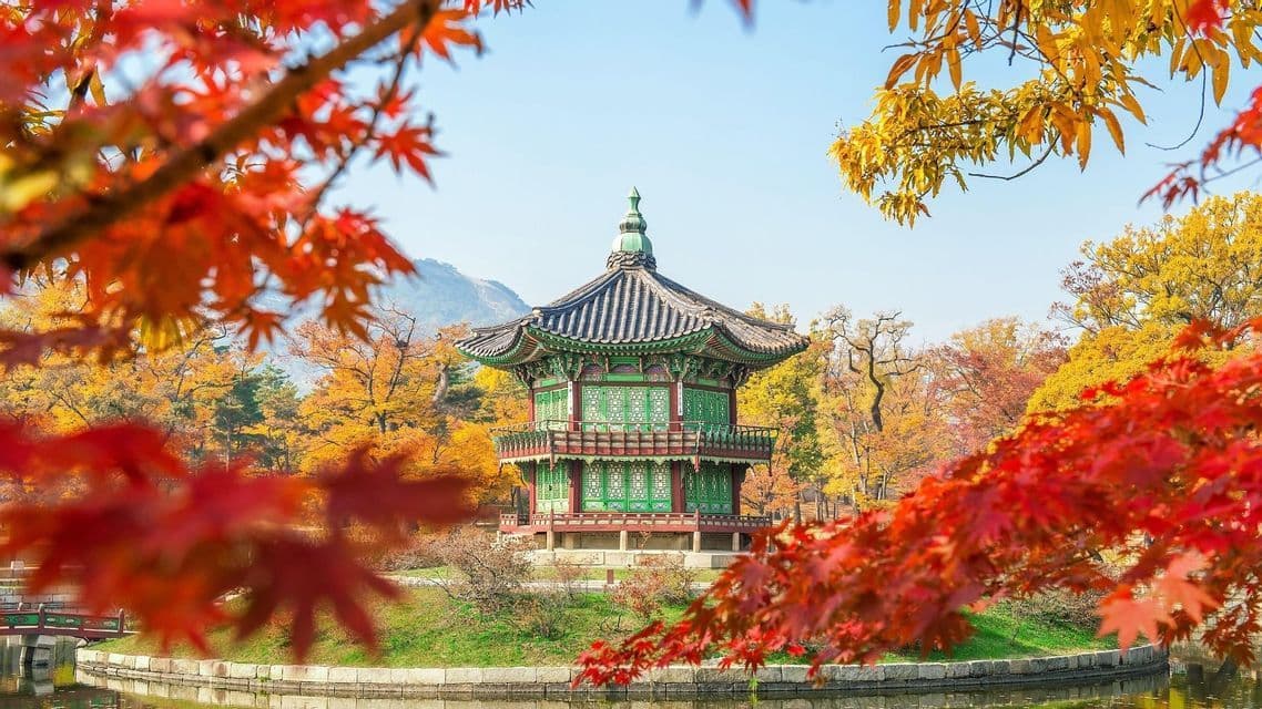 Ein traditioneller grün-roter Pavillon steht auf einer kleinen Insel in einem Teich, umgeben von farbenfrohem Herbstlaub.