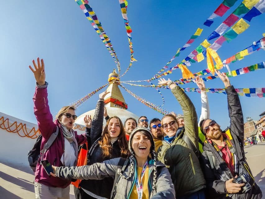 Un groupe WeRoad en voyage prend un selfie devant un grand stūpa, avec des drapeaux de prières colorés flottant dans le ciel bleu.