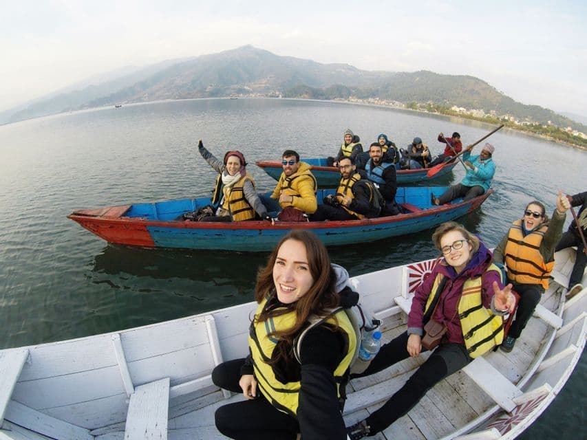 Un groupe WeRoad sourit pour un selfie alors qu'il navigue en bateaux colorés sur un grand lac, avec des montagnes brumeuses en arrière-plan.