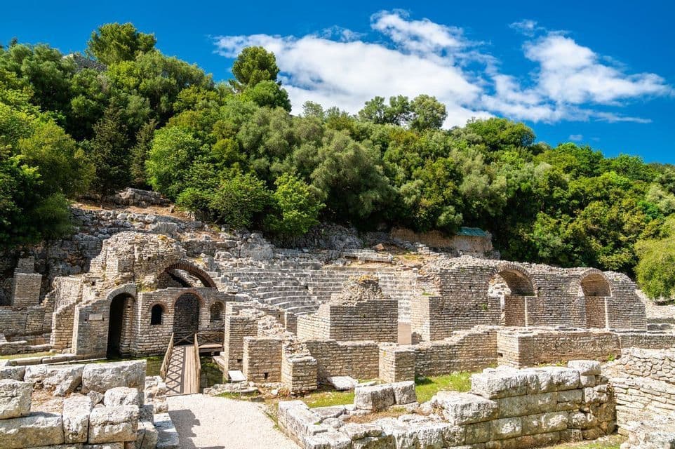 Antiguas ruinas de piedra de un anfiteatro anidado en una ladera cubierta de árboles verdes bajo un cielo azul.