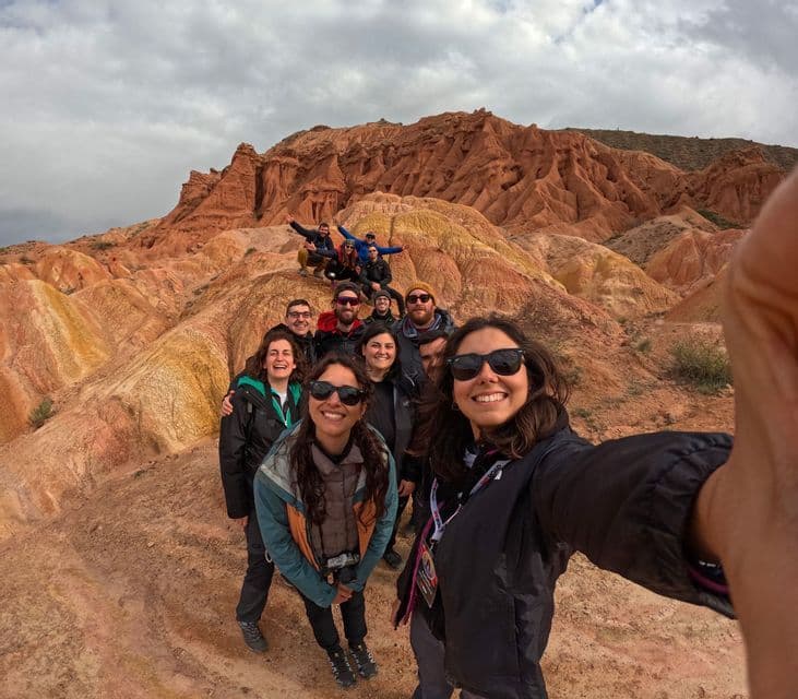 Eine lächelnde Frau macht ein Selfie mit ihrer WeRoad-Gruppe vor farbenfrohen roten und gelben Felsformationen unter einem bewölkten Himmel.