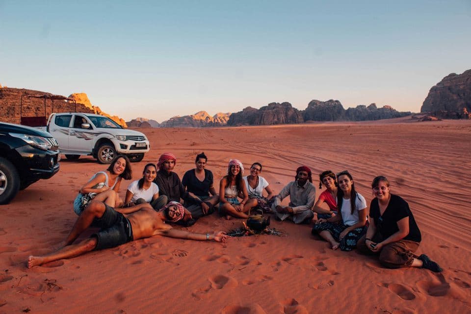 Un grupo de WeRoad, de viaje, se sienta en círculo sobre la arena roja de un desierto, alrededor de una tetera, con montañas de fondo al atardecer.