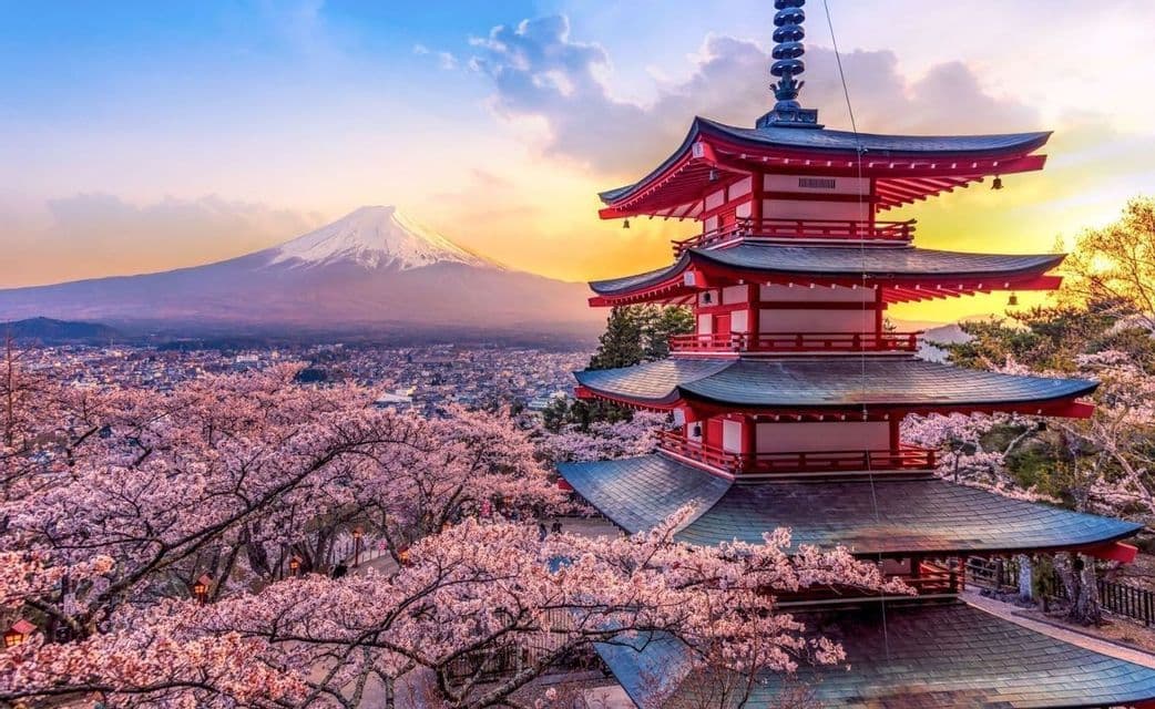 Una pagoda rossa si erge tra i fiori di ciliegio rosa, affacciata su una città con una montagna innevata sullo sfondo al tramonto.