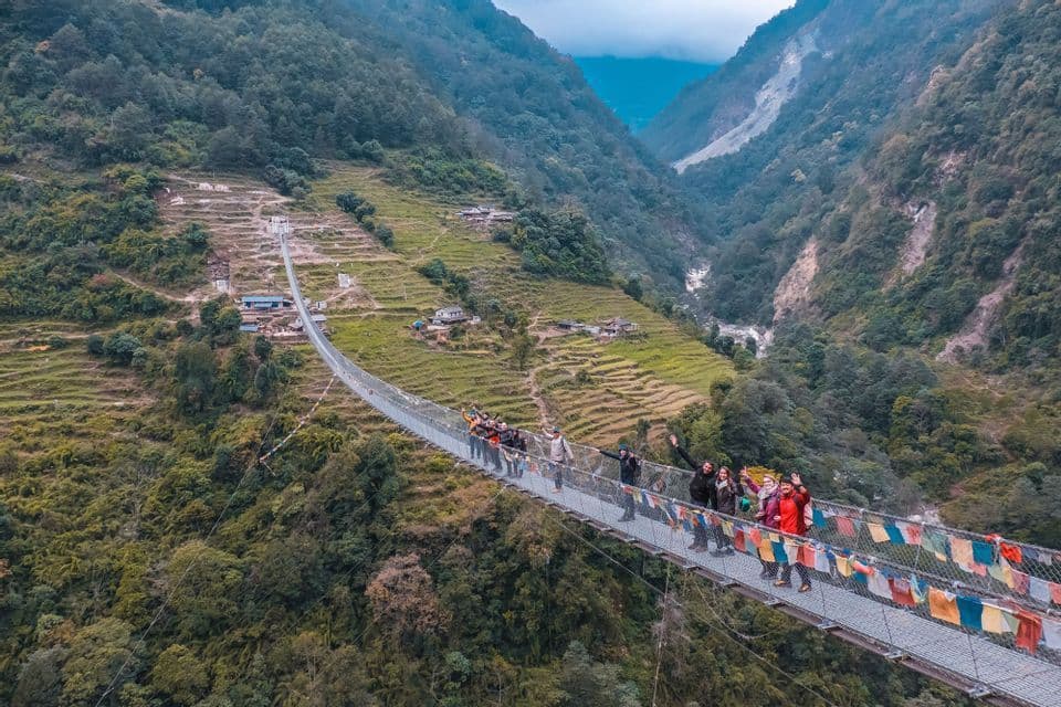Un grupo de WeRoad saluda desde un largo puente colgante, adornado con banderas de oración, que atraviesa un profundo valle verde.
