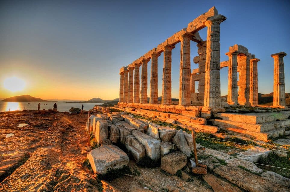 Antiguas ruinas de templos con grandes columnas de piedra en un acantilado rocoso, iluminadas por el sol poniente sobre el mar.