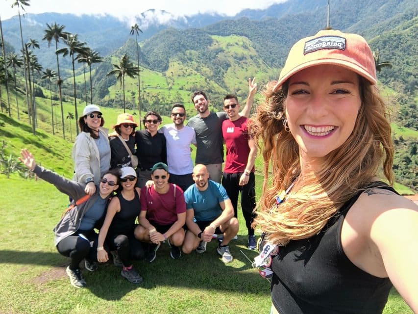 Un gruppo WeRoad in viaggio si scatta un selfie su una collina verde con alte palme e montagne sullo sfondo.