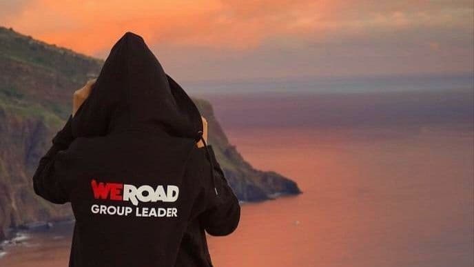 Un Group Leader WeRoad con una felpa nera osserva un tramonto sull'acqua accanto a una scogliera.