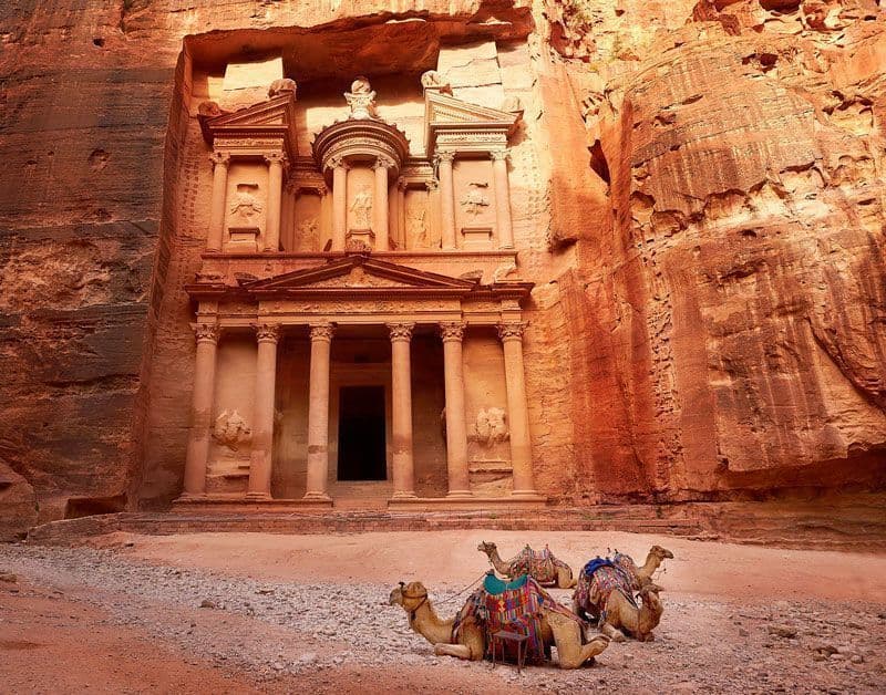 Trois chameaux aux selles colorées se reposent au pied d'une imposante façade de temple taillée dans une falaise de roche rouge.