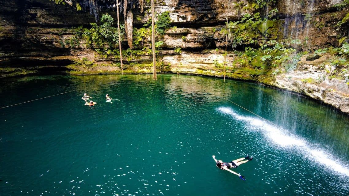 An aerial view of a WeRoad group trip swimming and floating in the turquoise water of a cenote surrounded by rocky cliffs.