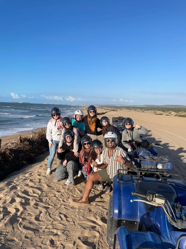Eine WeRoad-Gruppe mit Helmen posiert mit ihren Quads auf Sanddünen vor dem Meer.