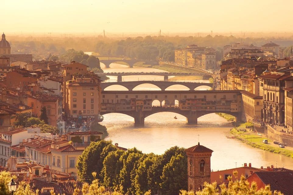 Una vista aérea de una ciudad histórica con puentes arqueados cruzando un río ancho bajo un cielo brumoso y dorado.