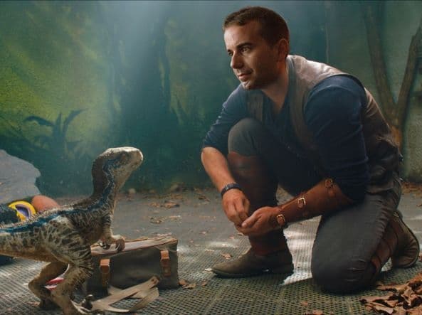 Un hombre con chaleco se arrodilla en un suelo metálico, mirando a un pequeño dinosaurio de rayas azules de pie sobre una bolsa de lona.