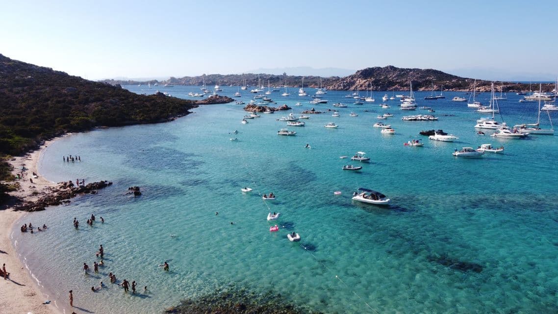 Barche e yacht all'ancora in una bassa baia turchese con persone che nuotano vicino alla riva sabbiosa.