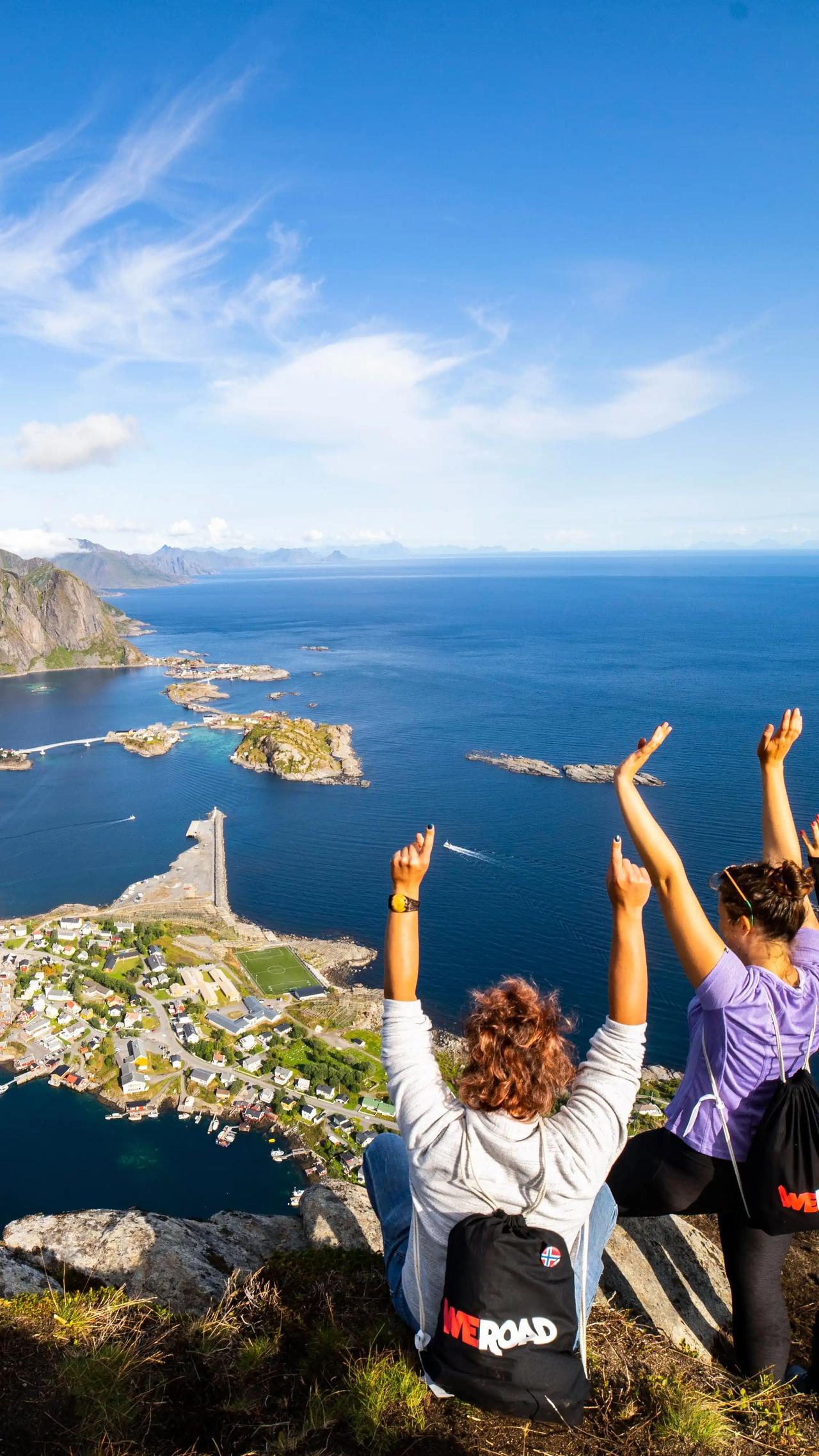 Deux voyageurs WeRoad les bras levés au sommet du Reinebringen aux îles Lofoten, avec une vue panoramique sur le village de pêcheurs de Reine, les îles et le fjord bleu profond en contrebas