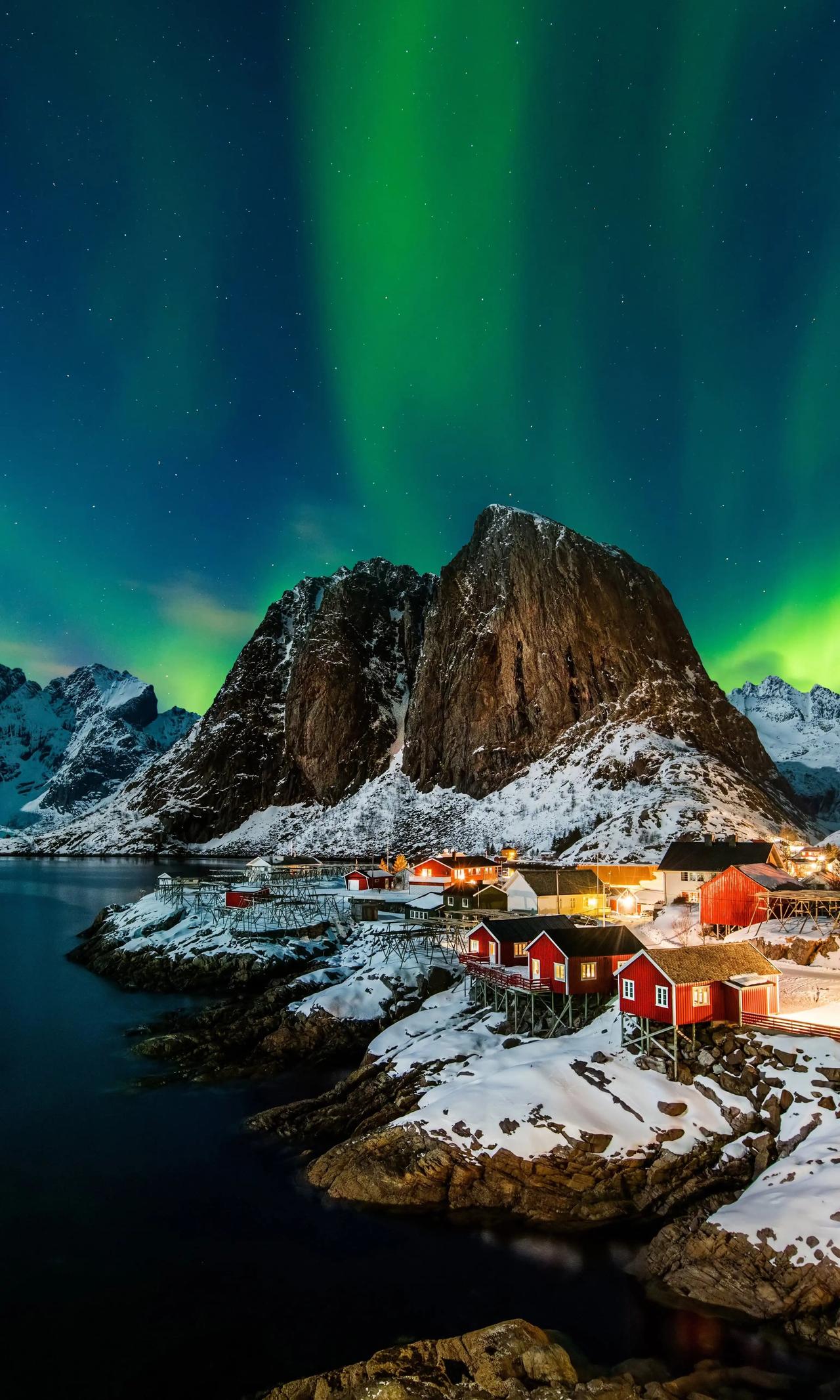Aurores boréales vertes au-dessus du village de pêcheurs enneigé de Hamnøy aux îles Lofoten en Norvège, avec des maisons en bois rouges sur pilotis, un fjord calme et un pic rocheux imposant en arrière-plan