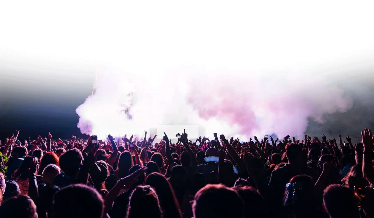 Público con los brazos en alto frente al escenario de un concierto con humo y luces.