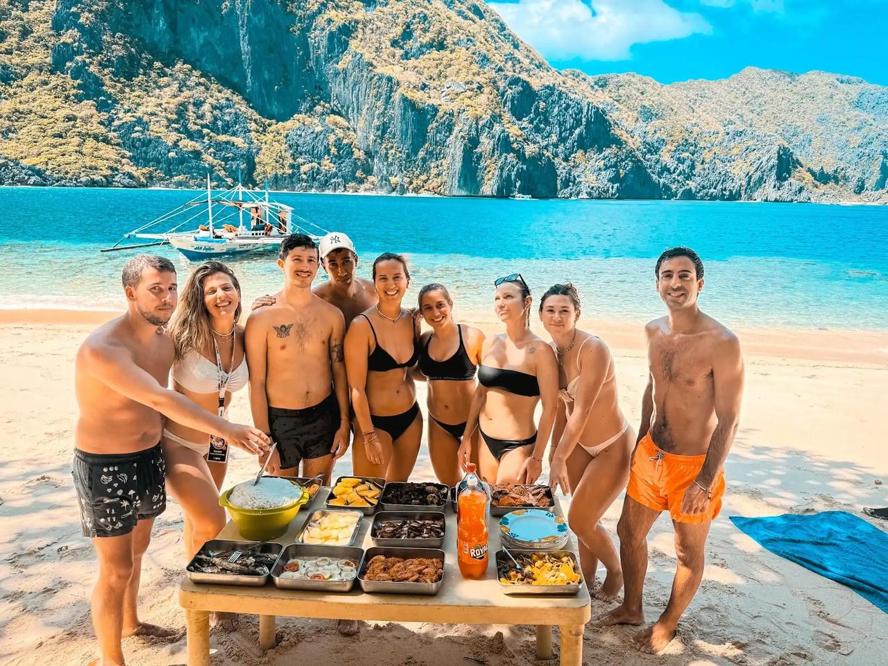 Un grupo de WeRoad en bañador posa en la playa junto a una mesa con comida, con acantilados y un barco al fondo.