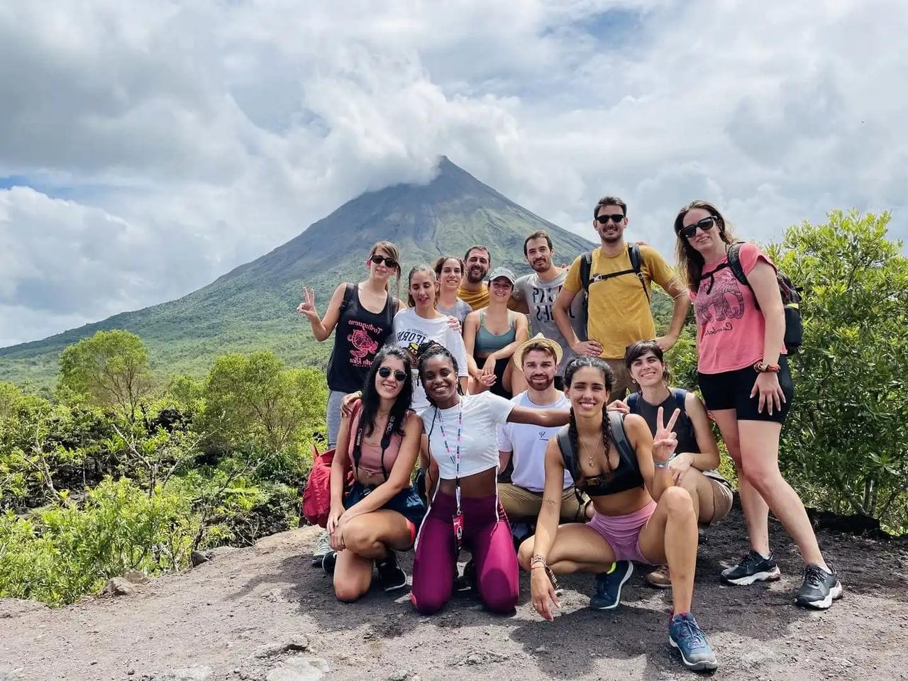 Eine WeRoad-Gruppe posiert auf einem Wanderweg für ein Gruppenfoto, im Hintergrund ein großer, wolkenverhangener Vulkan.