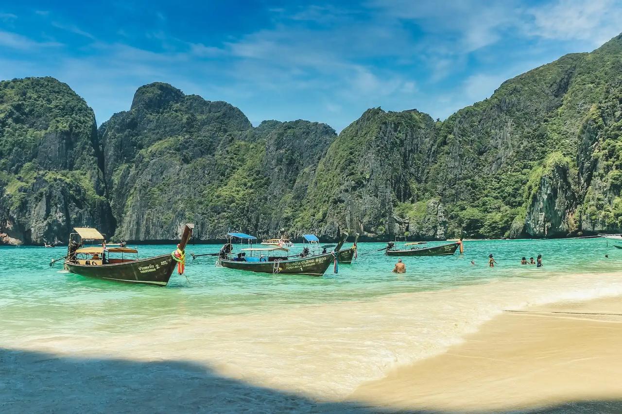 Bateaux longtail traditionnels amarrés dans la baie de Maya Bay sur l'île de Koh Phi Phi avec mer turquoise, plage de sable blanc et imposantes falaises calcaires couvertes de végétation, Thaïlande