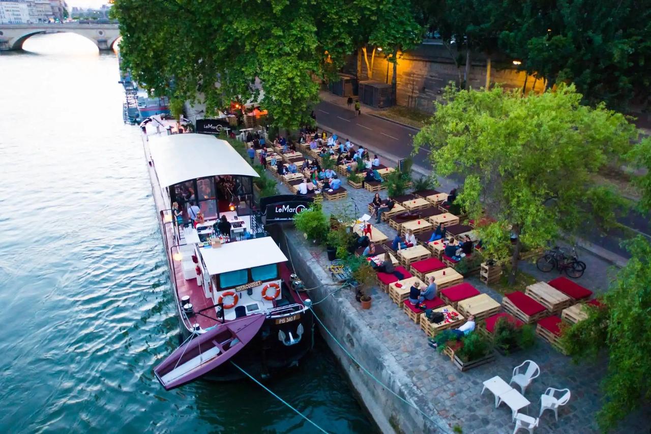 Barco-restaurante amarrado junto a terrazas llenas de gente a la orilla de un río en la ciudad.