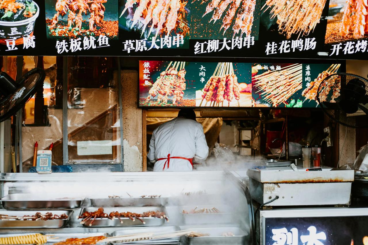 Un cuoco lavora in una bancarella fumante di street food, con spiedini in mostra sotto un menu luminoso e caratteri cinesi.
