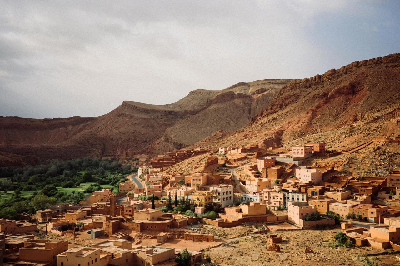 Un pueblo de edificios de tonos tierra, construido en una ladera montañosa rocosa y roja, con un valle verde a la izquierda bajo un cielo nublado.