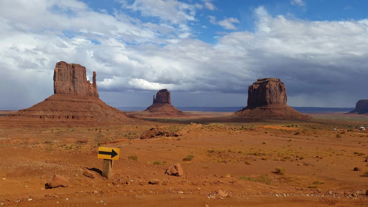 Large red rock buttes stand in a vast desert under a cloudy sky, with a yellow arrow sign in the foreground.