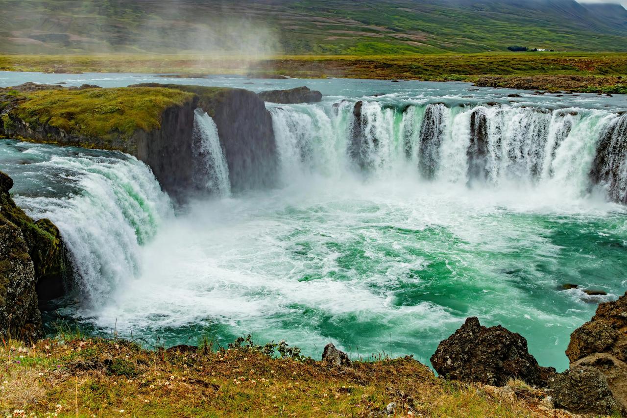 Ein breiter, mächtiger Wasserfall stürzt über moosbewachsene Klippen in einen turbulenten türkisen Fluss unter einem bewölkten Himmel.
