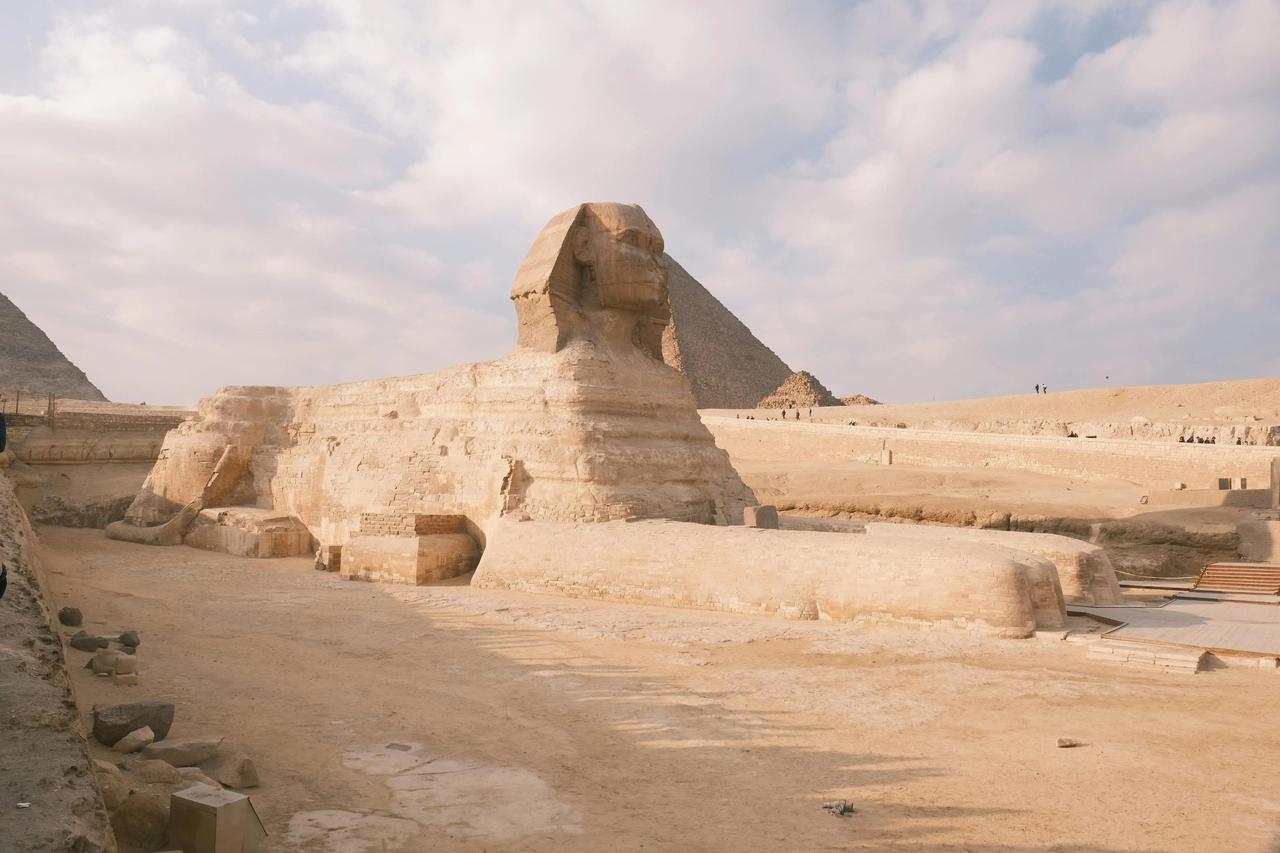 El monumento de la Gran Esfinge descansa en un paisaje arenoso con una gran pirámide visible al fondo bajo un cielo parcialmente nublado.
