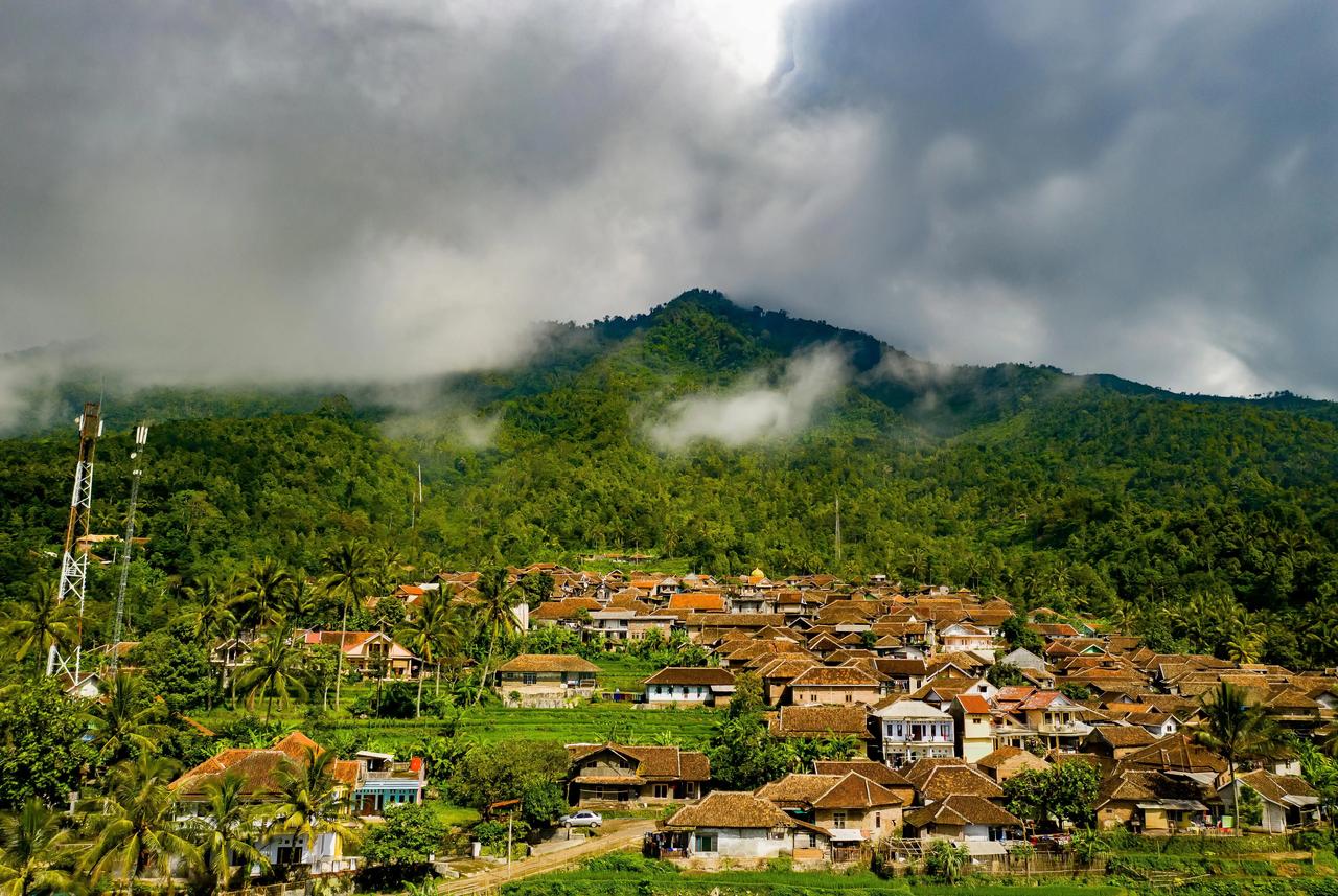 Vue aérienne d'un village aux toits de tuiles brunes, niché au pied d'une montagne verdoyante et brumeuse.