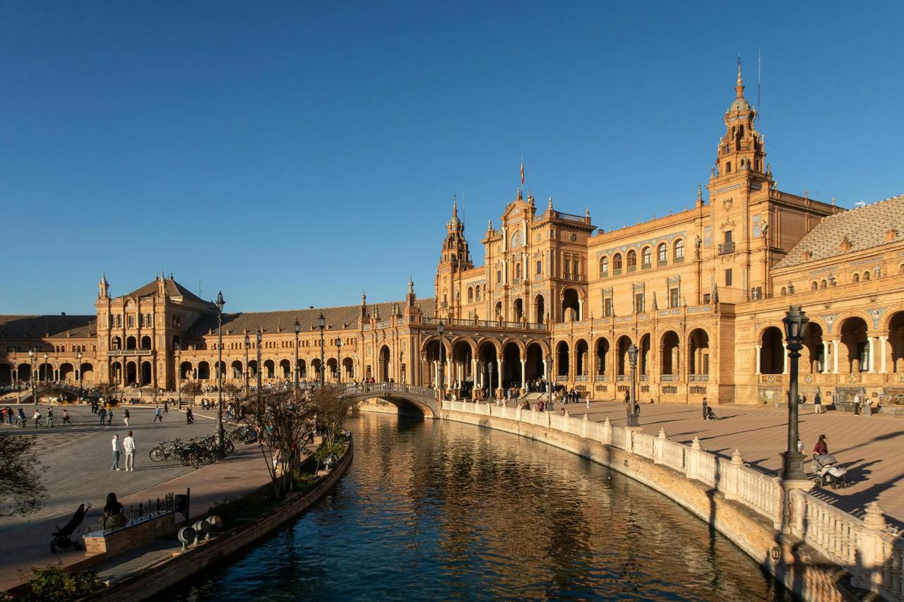 Un bâtiment historique et orné, avec des arches et des tours, borde un canal sinueux, des gens se promenant sur la place sous un ciel d'un bleu limpide.