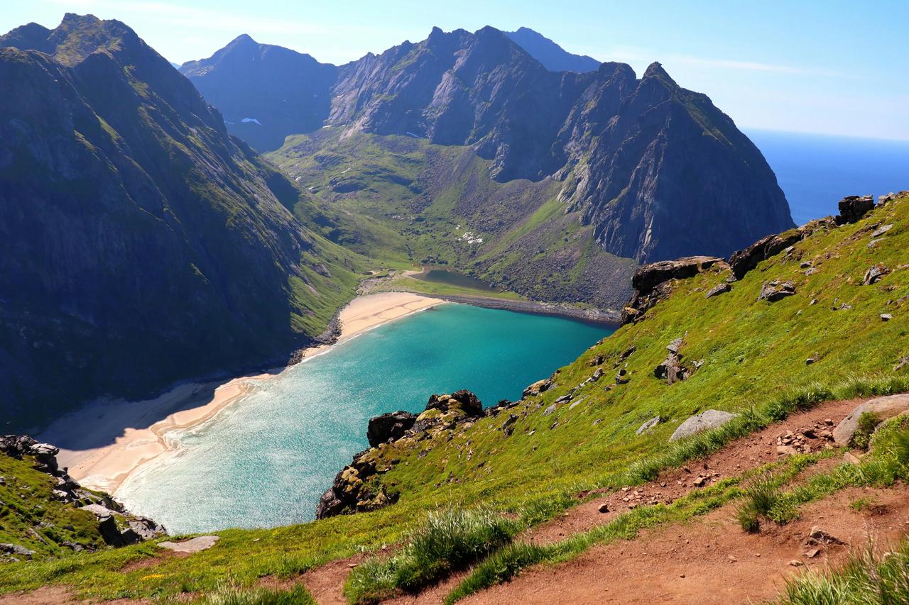 Una vista da un pendio montano erboso con un sentiero escursionistico si affaccia su una caletta turchese e una spiaggia sabbiosa circondata da ripide cime.