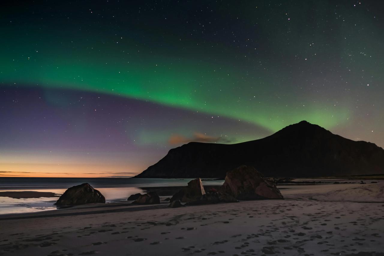 L'aurora boreale verde risplende in un cielo notturno stellato sopra una spiaggia sabbiosa con grandi rocce e la sagoma di una montagna.