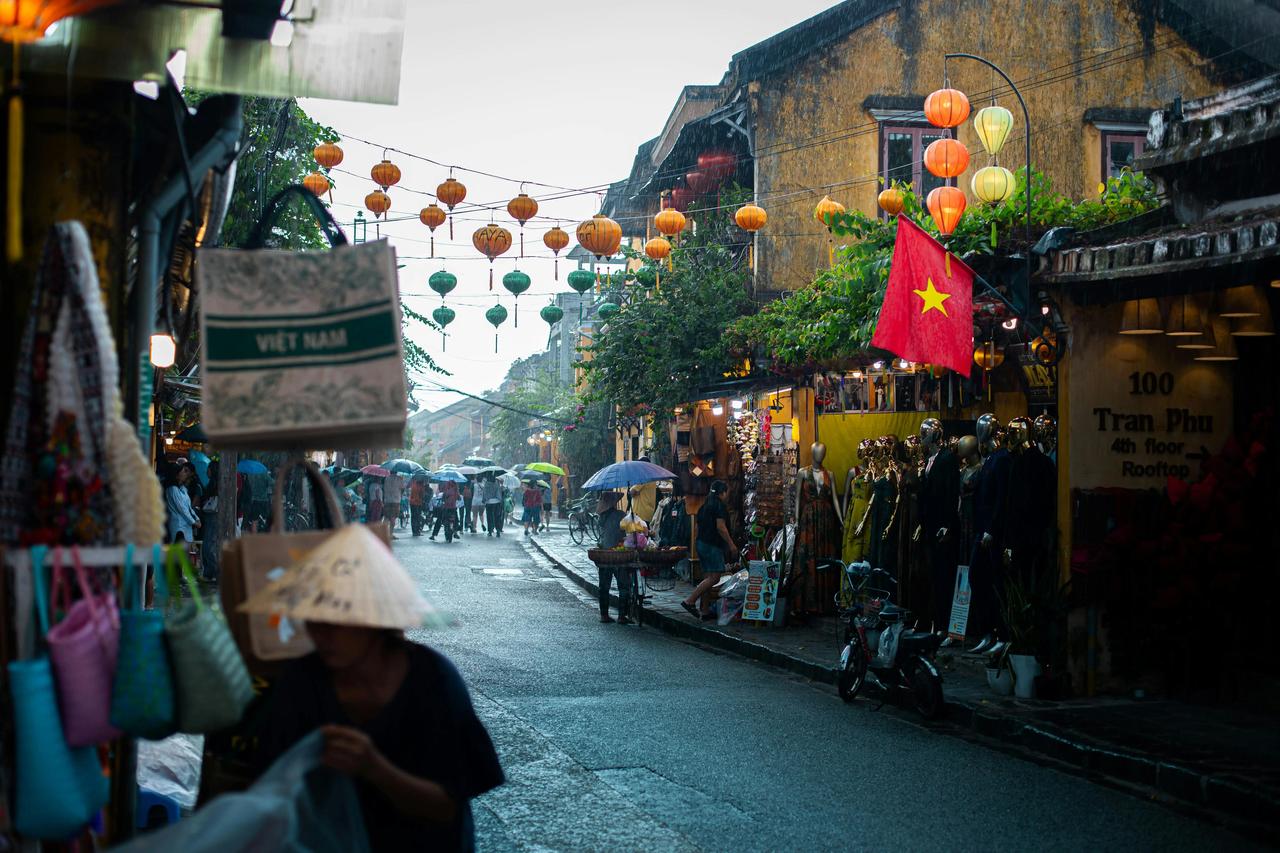 Des passants sous des parapluies déambulent dans une rue vietnamienne pluvieuse, bordée de boutiques et ornée de lanternes en papier colorées.