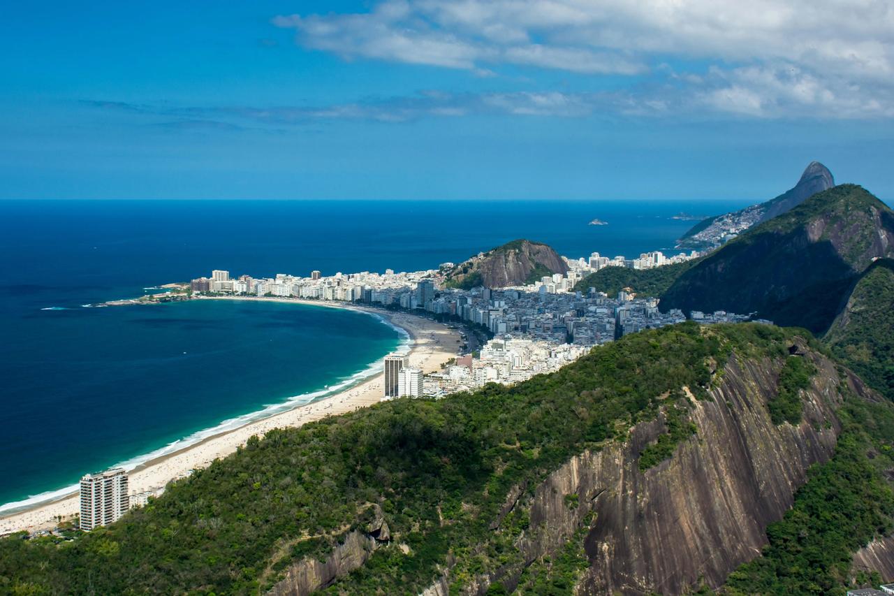 An aerial view of a coastal city along a curved sandy beach, nestled between green mountains and the blue ocean.