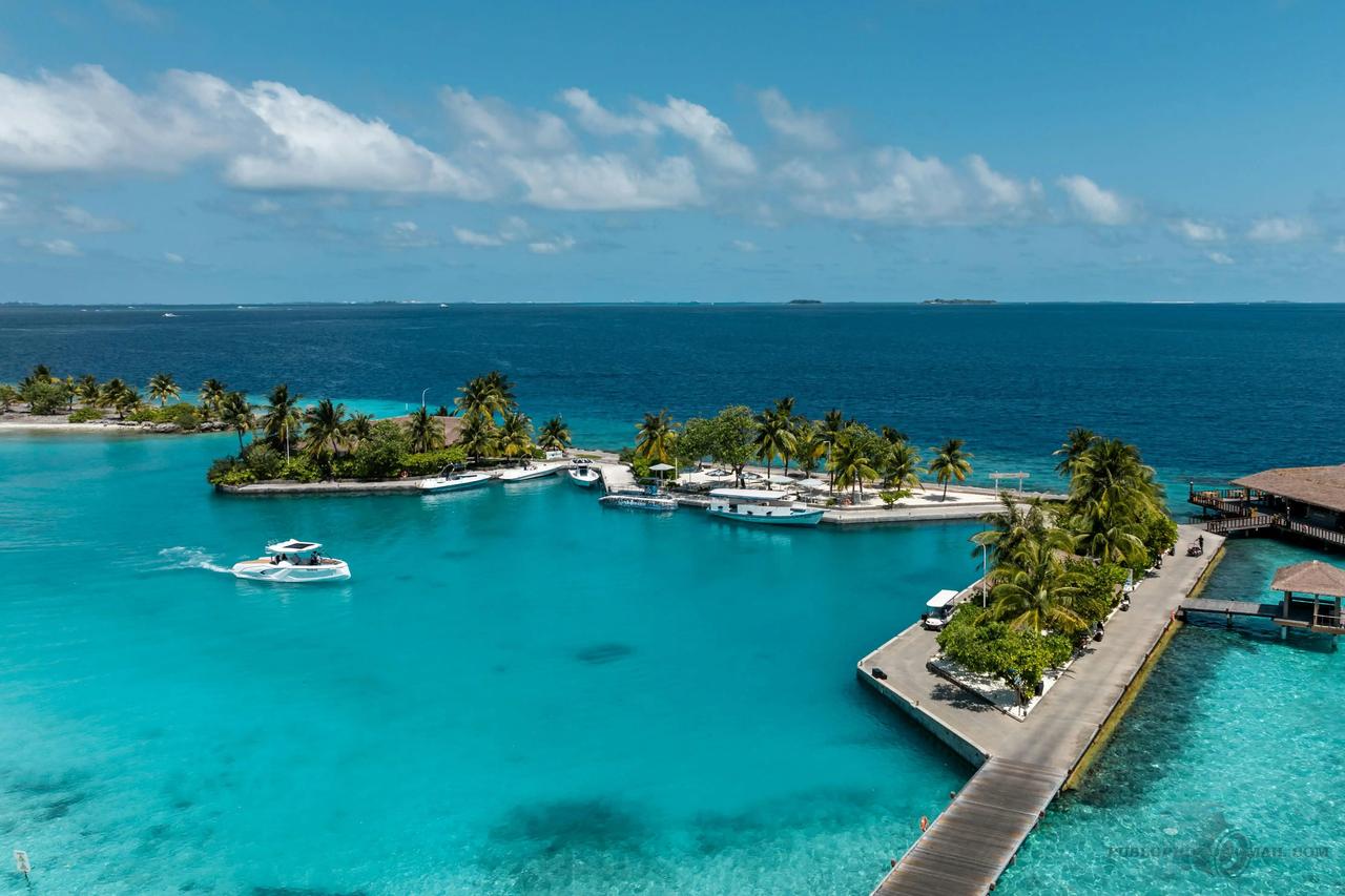 Vista aerea di un motoscafo su acqua turchese vicino a una piccola isola tropicale con un molo e palme.