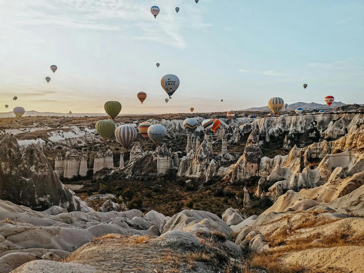 Decenas de globos aerostáticos flotan sobre un valle rocoso con formaciones cónicas bajo un cielo brumoso al amanecer.