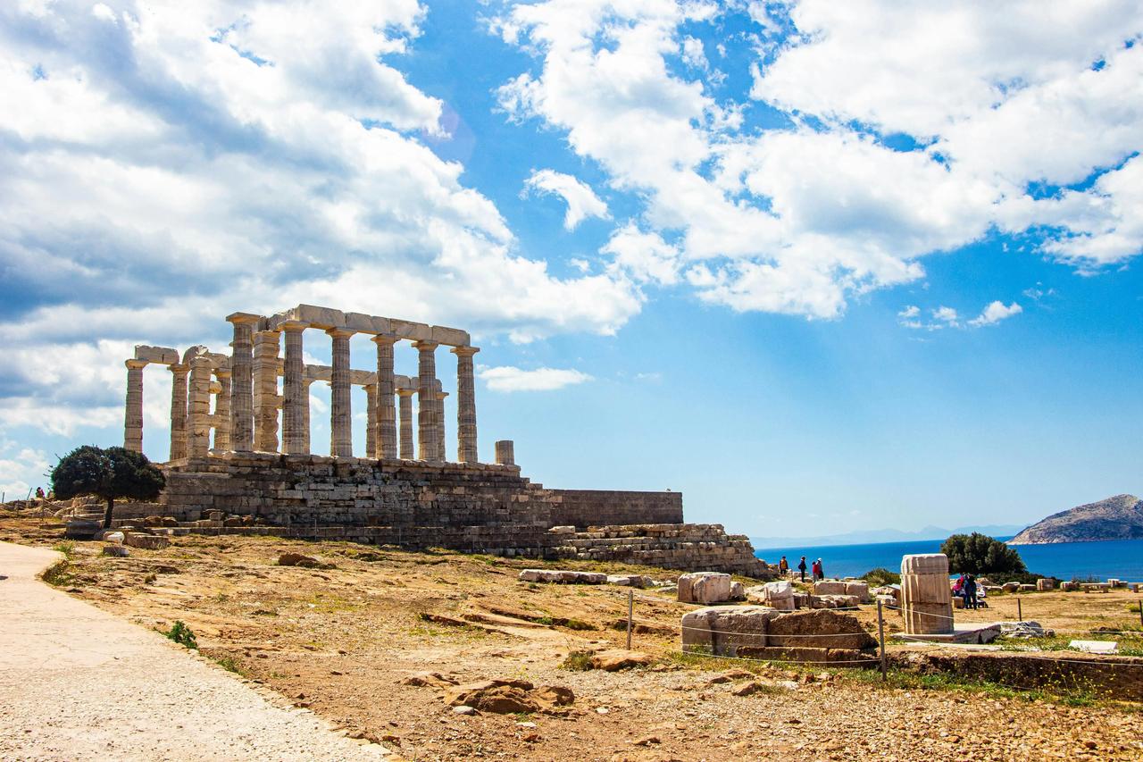 Le rovine di un antico tempio con grandi colonne si ergono su un promontorio roccioso che domina il mare sotto un cielo parzialmente nuvoloso.