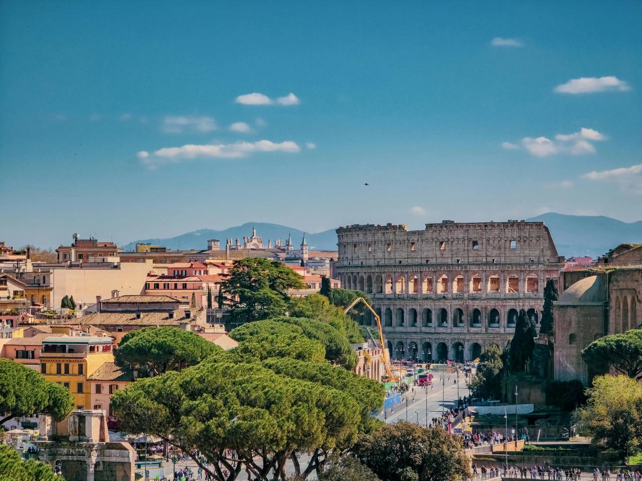 Un paesaggio urbano diurno con il Colosseo, circondato da edifici storici e alberi verdi lussureggianti sotto un cielo azzurro.