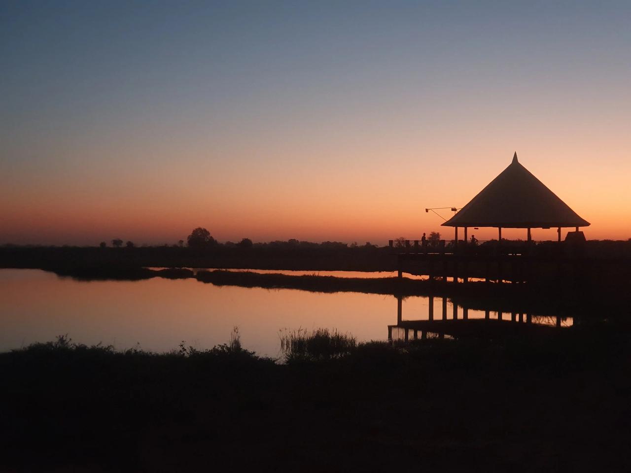 Silueta de una cabaña con techo de paja junto a un lago al atardecer.